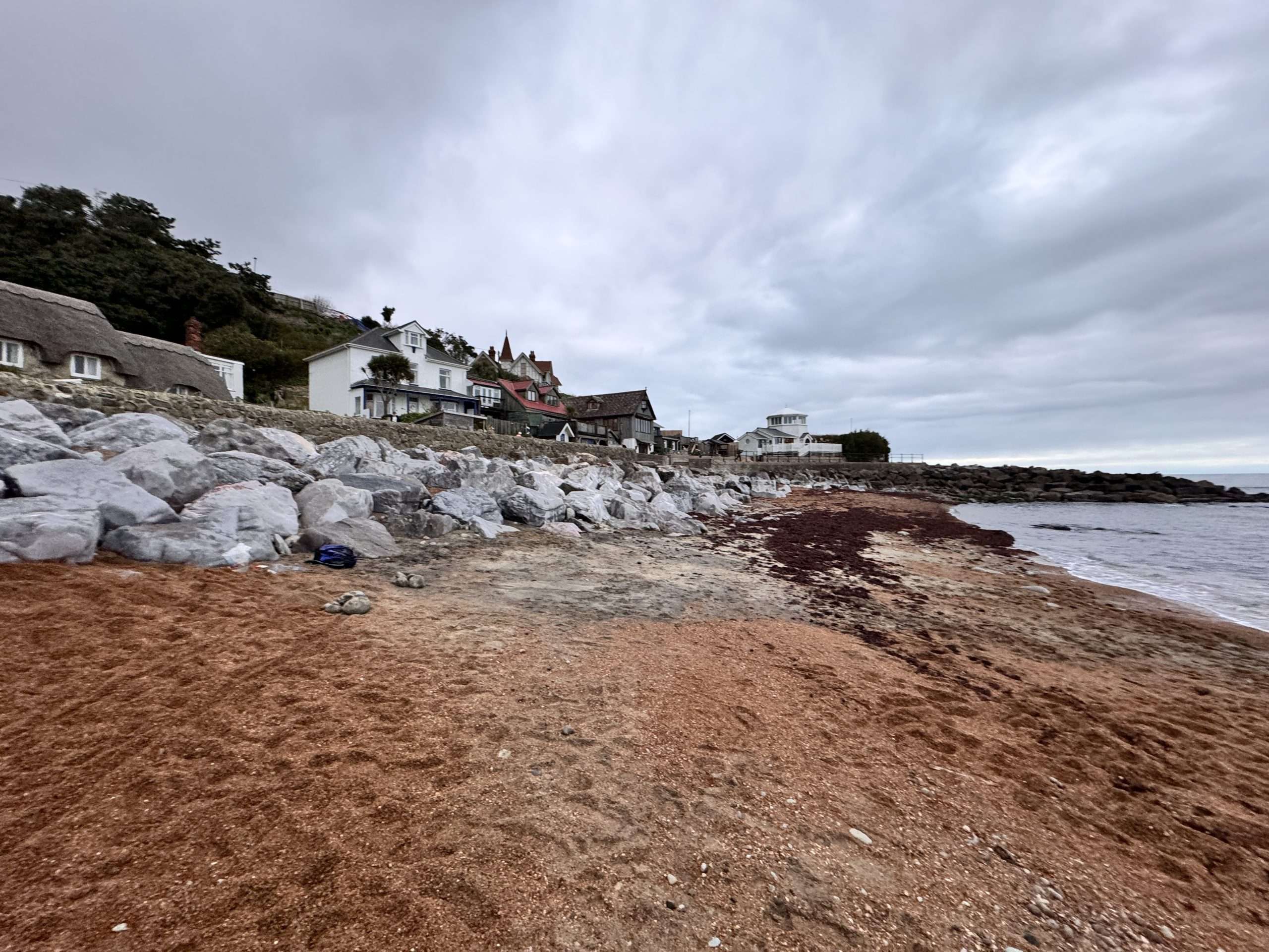 Coastal village with homes protected by a rock sea defence along an eroding beach under overcast skies, highlighting shoreline management and flood risk planning.